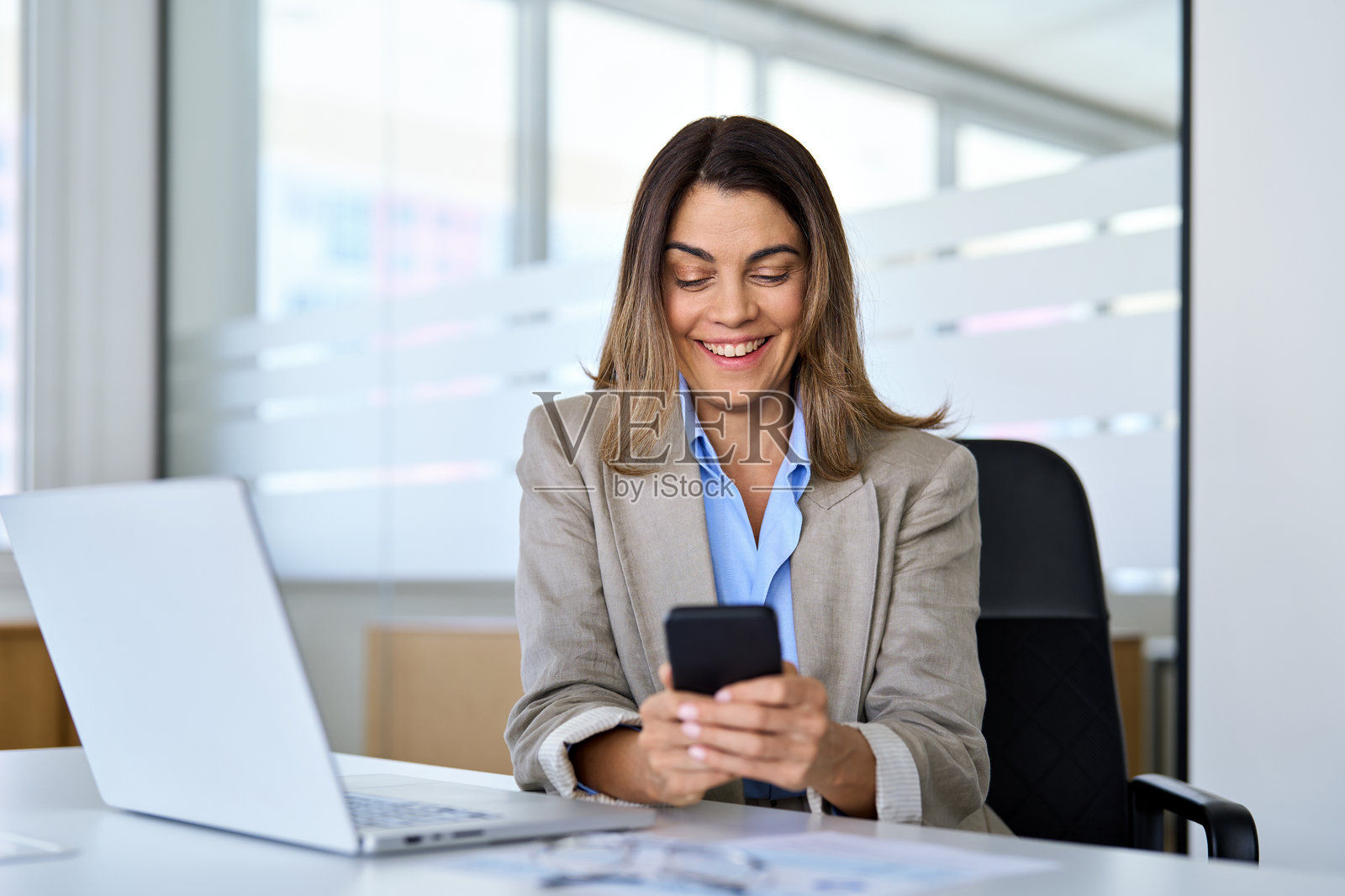 快乐而忙碌的中年女商人正在办公室使用智能手机。照片摄影图片