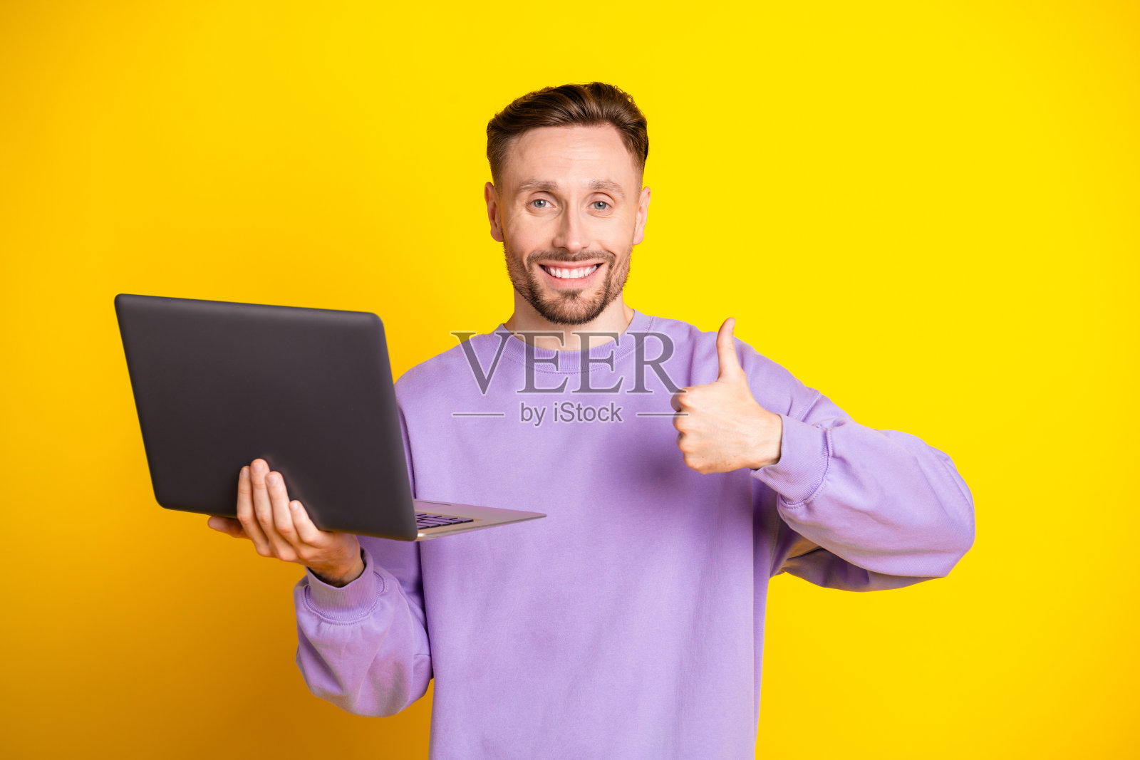 年轻男子手持笔记本电脑,面带微笑,竖起大拇指,背景为明亮的黄色照片摄影图片