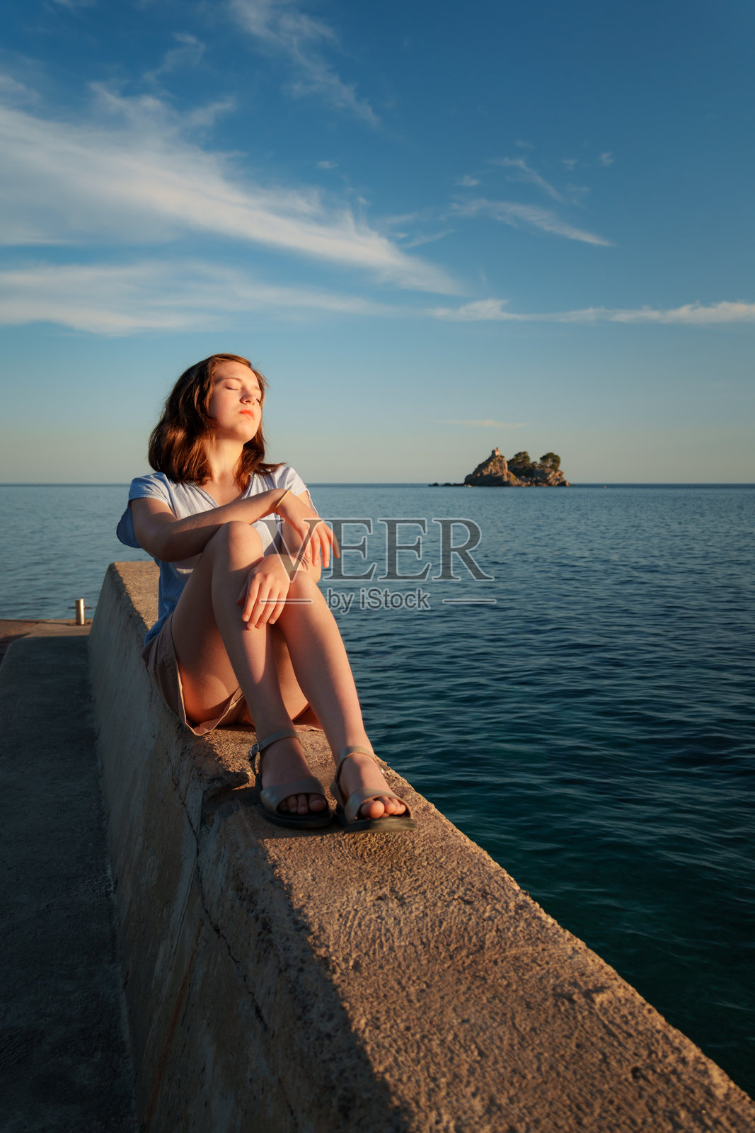 在黑山佩特罗瓦茨,年轻女子在日落时分放松地坐在石码头上。她闭着眼睛,这是一个全身镜头,表情安详,充满了夏日假期和海风的惬意。照片摄影图片