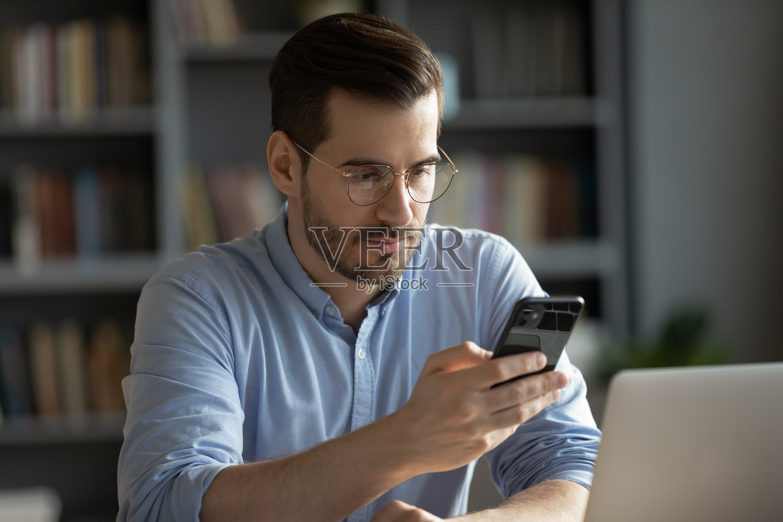 年轻的商务人士坐在书桌旁,一边看着笔记本一边使用智能手机照片摄影图片