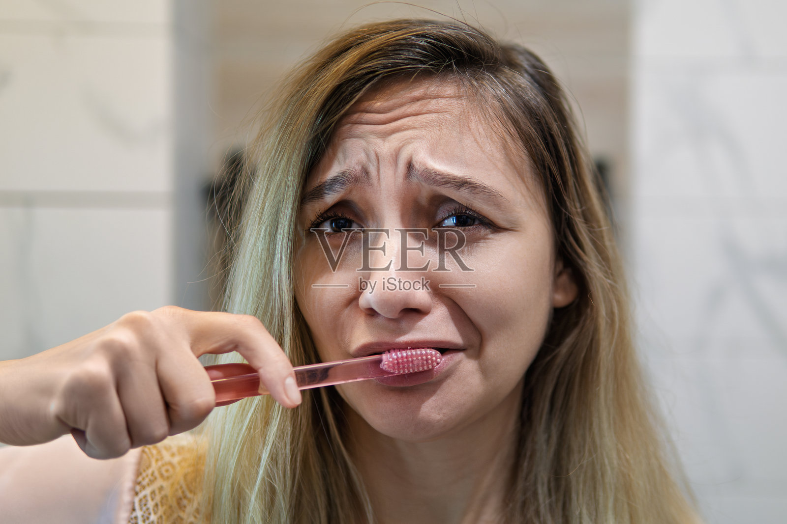 一个悲伤的女孩嘴里含着牙刷,看着镜子。牙痛和痛苦的概念。一位年轻女子正在刷牙。照片摄影图片