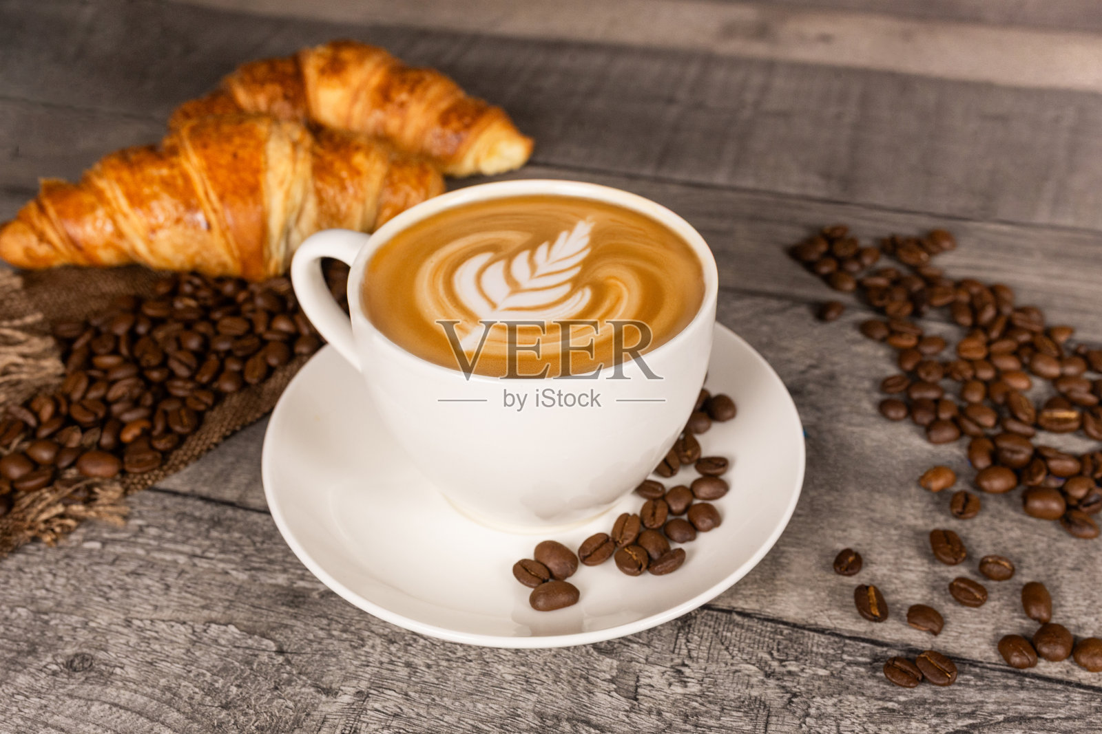 木质背景上的卡布奇诺咖啡,带有拉花艺术、羊角面包和咖啡豆。照片摄影图片