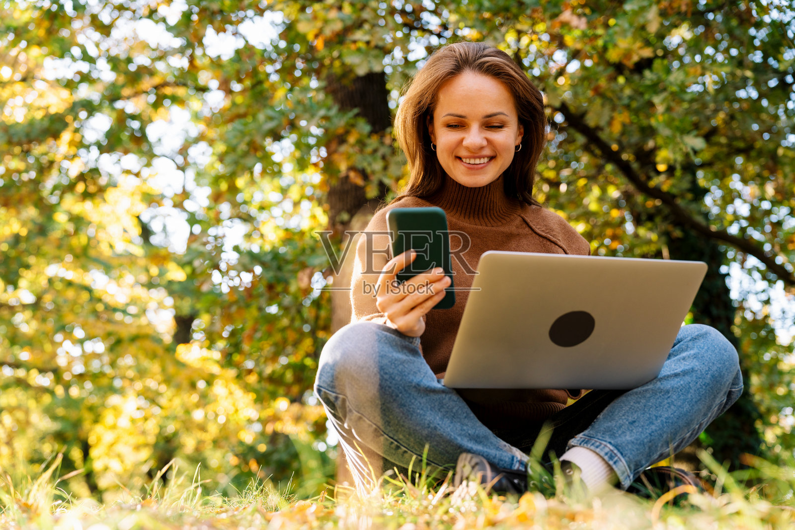 一位现代女性坐在公园的地上,同时使用智能手机和笔记本电脑。照片摄影图片