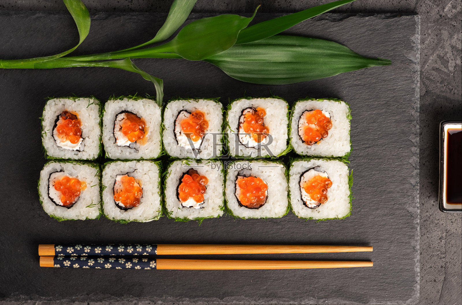 新鲜的寿司卷整齐地摆放在深色石板盘上,饰以绿叶,配有筷子和酱油,展现出日式料理充满活力的色彩和质感照片摄影图片