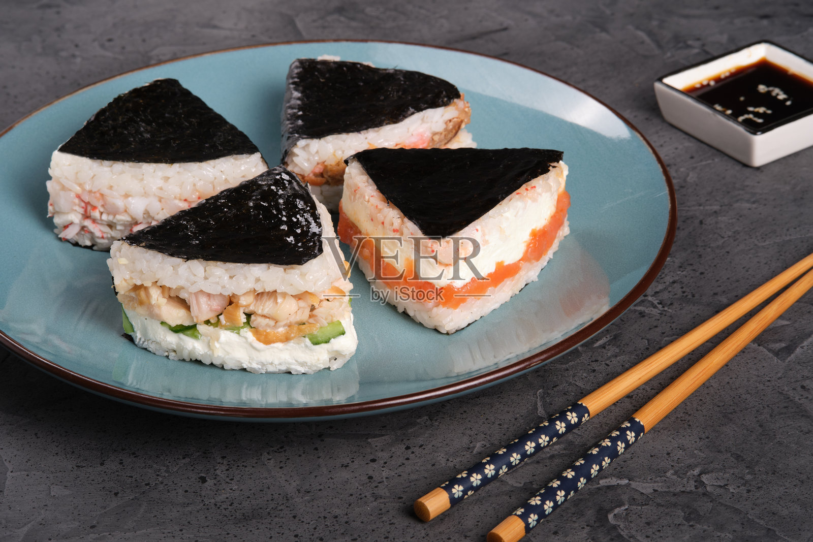 蓝盘上摆放着色彩鲜艳的寿司三角,内含多种馅料,并饰有海苔,配有筷子和酱油,带来愉悦的美食体验照片摄影图片