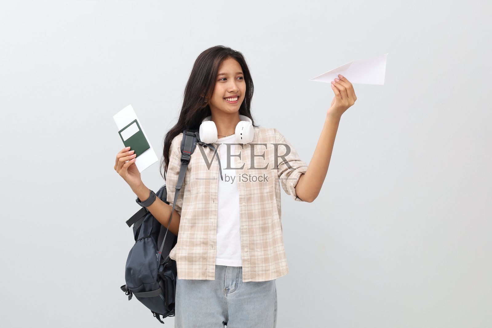 年轻女学生穿着休闲服,背着背包,脖子上挂着耳机,手持护照和机票,旁边是飞机模型,呈现度假概念和航空旅行,孤立地站立在白色背景上。照片摄影图片