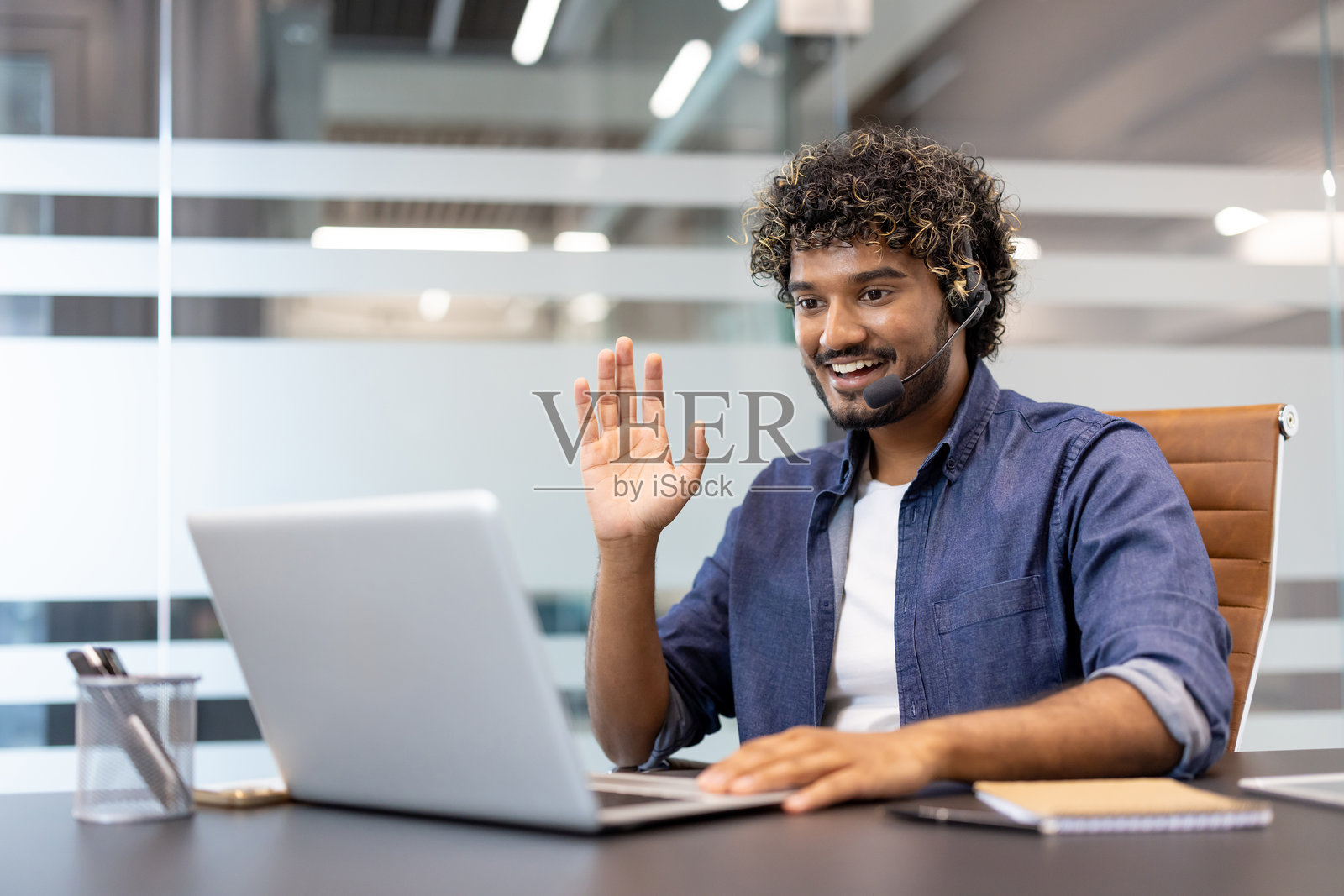 年轻的印度男子戴着耳机坐在办公室的桌子旁,在笔记本电脑上进行视频通话,并微笑着挥手致意照片摄影图片