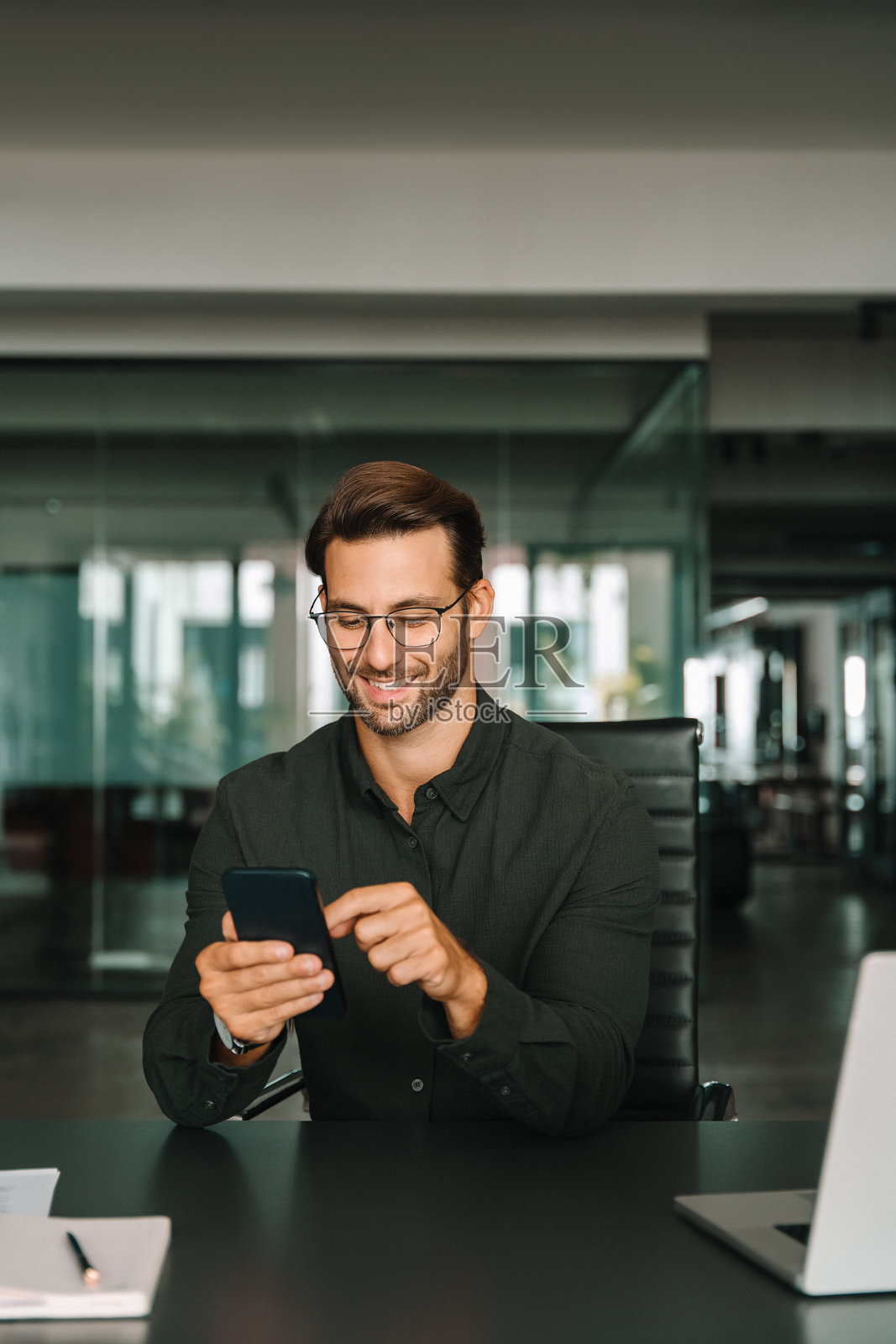 微笑的拉丁裔西班牙裔商务经理使用手机移动应用程序和笔记本电脑。欧洲年轻学生男商人手持智能手机坐在办公室里使用小工具在线工作。竖屏,可留白。照片摄影图片