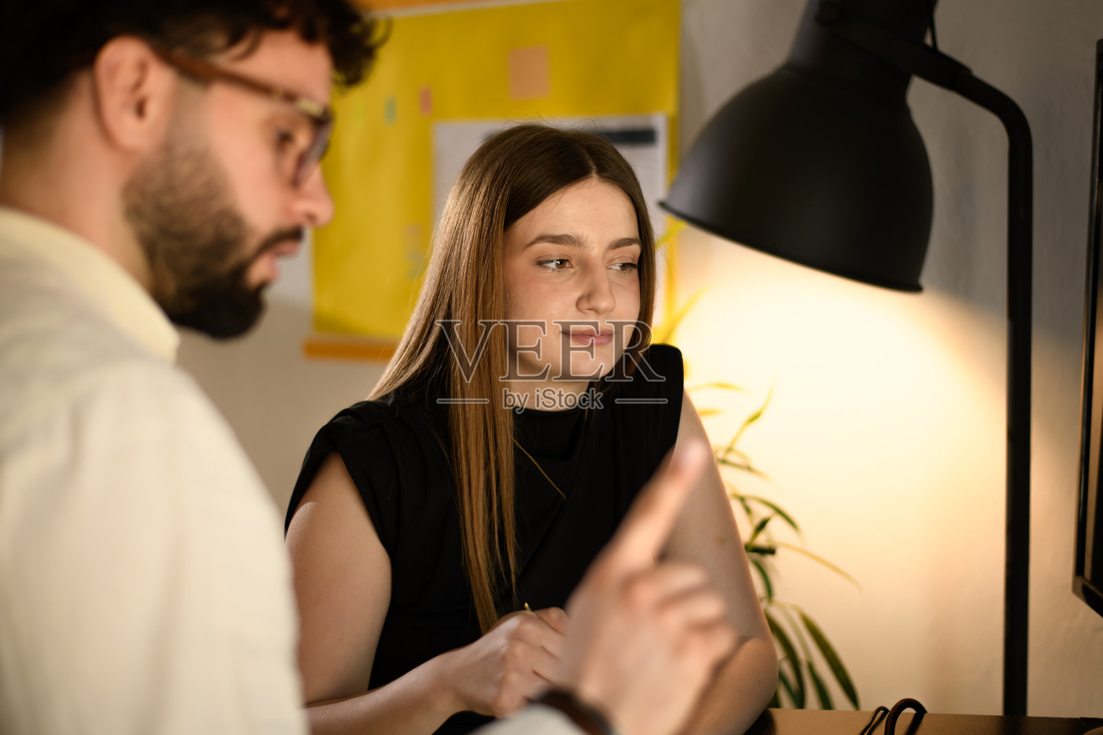 在办公室环境中,一位男商人与一位女士正在讨论项目,背景为暖色灯光和绿植。照片摄影图片