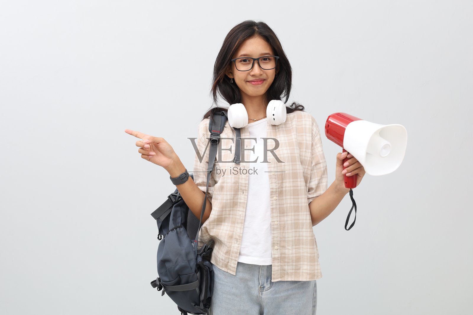 一位穿着休闲服、戴着耳机、背着背包的亚洲女学生,手持扩音器指向旁边空无一物的空间,脸上带着微笑看向镜头。照片摄影图片