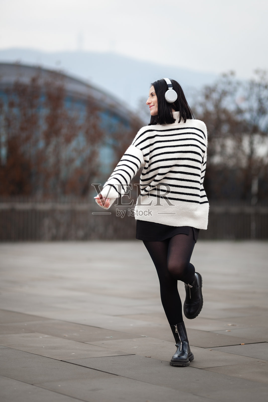 一个女孩在现代都市背景下,穿着冬装,戴着耳机,在户外快乐地跳舞,背景虚化照片摄影图片