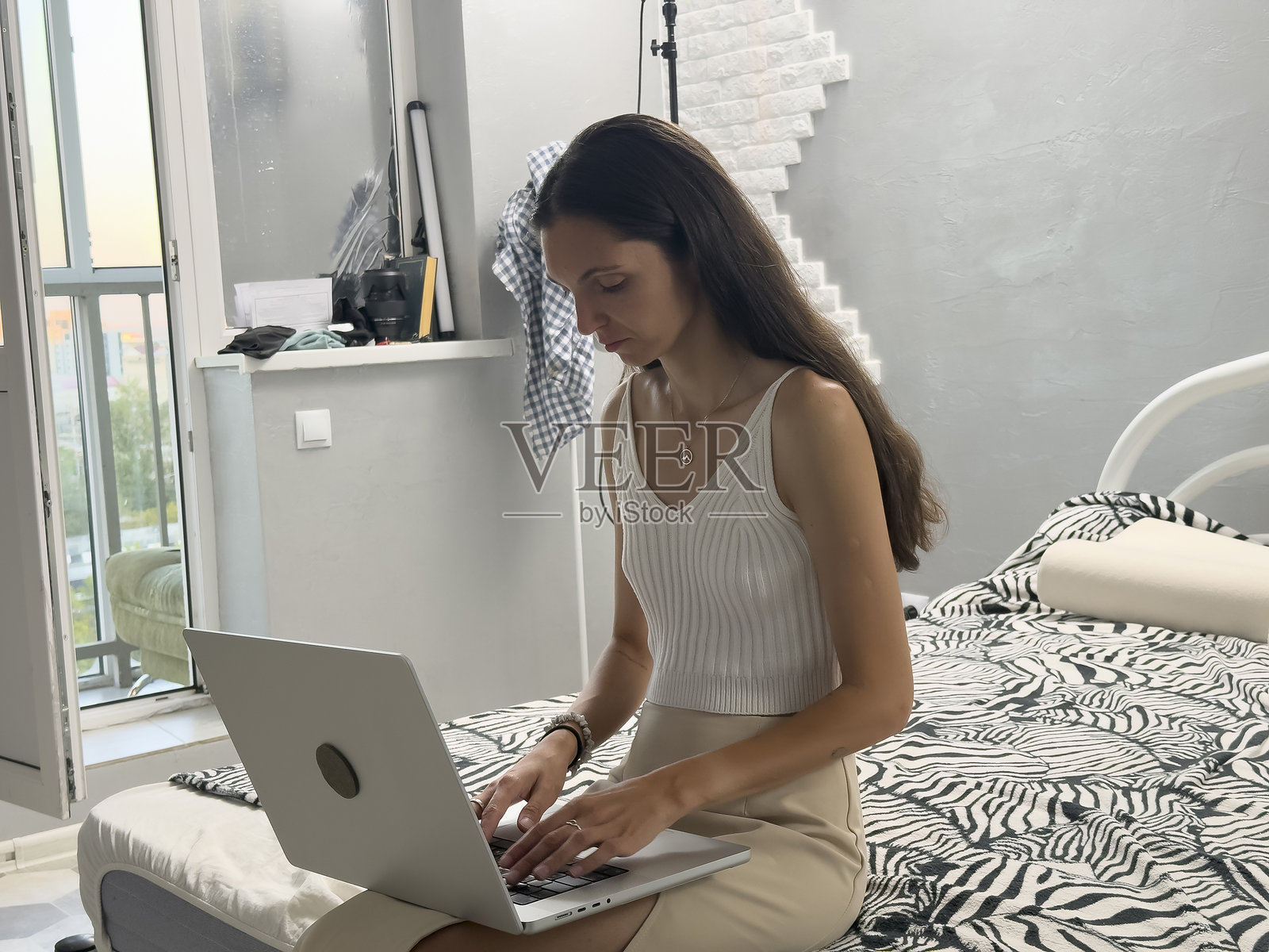 一个女人坐在床上,在一间家具稀疏的小房间里用笔记本电脑打字照片摄影图片