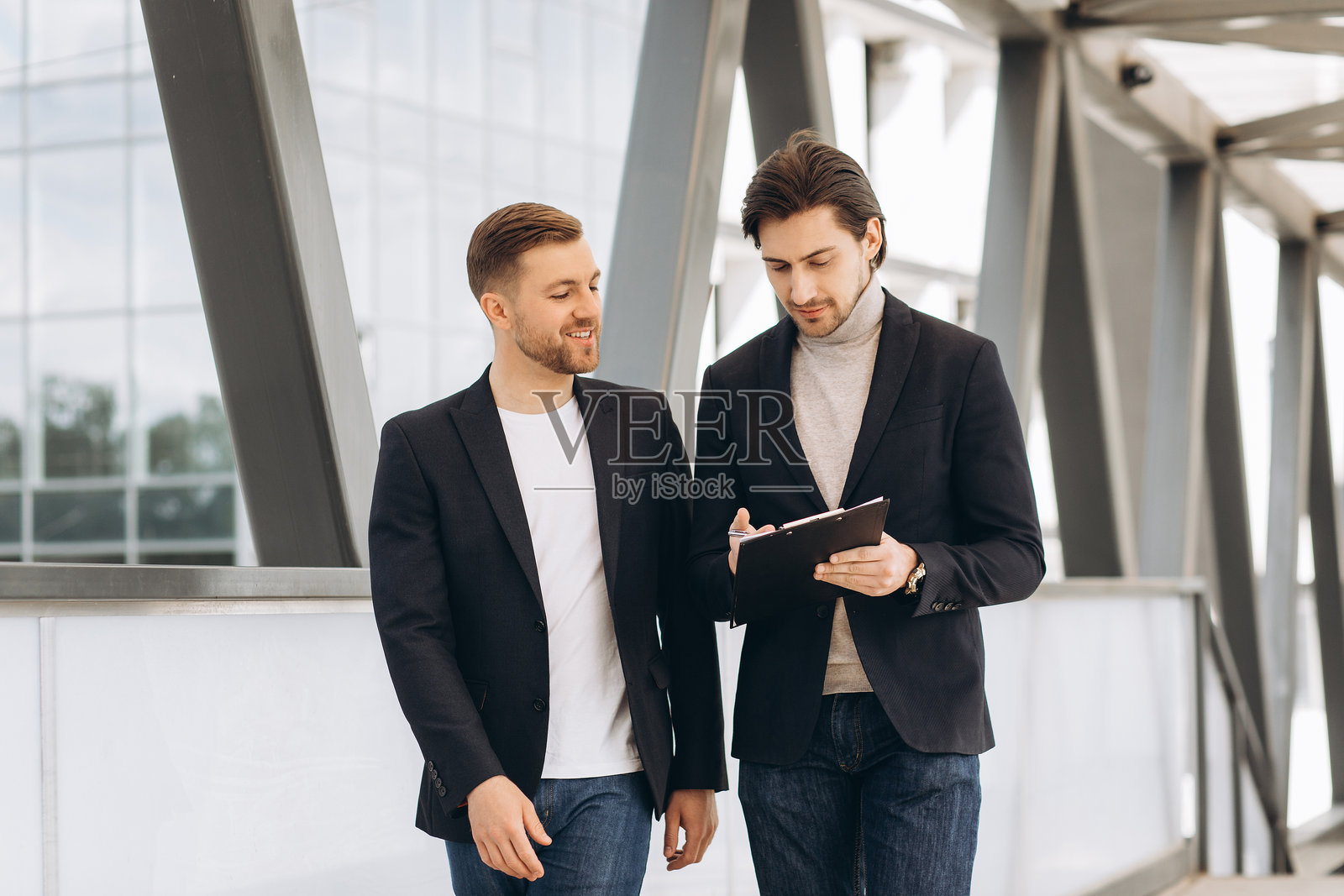 两位专业的商业伙伴在户外边走边讨论项目细节,并查看剪贴板上的文件。照片摄影图片