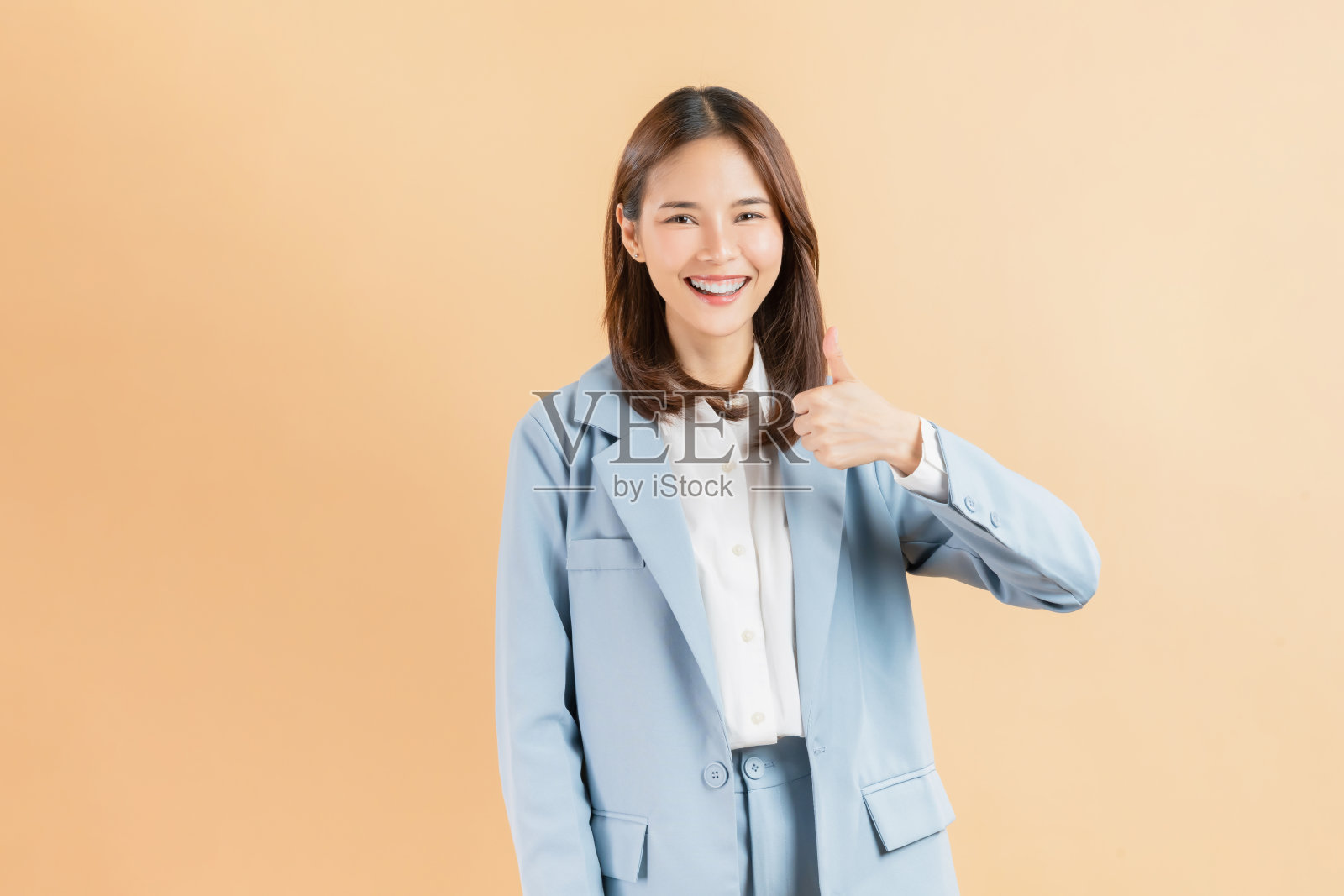 一位微笑的亚洲女商人热情地竖起大拇指,在米色背景上表示赞同或喜欢。照片摄影图片