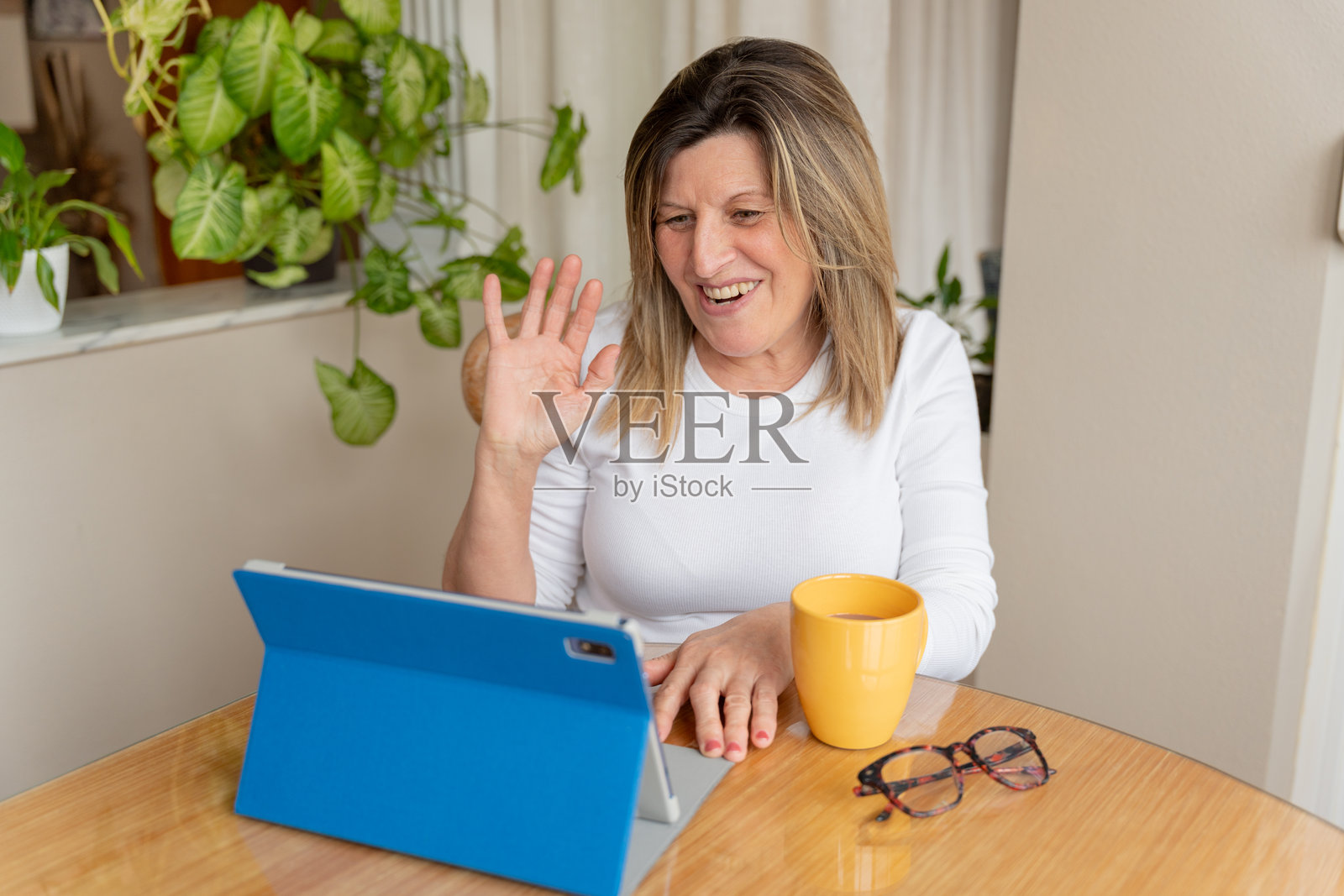 一位成熟的成年女性在视频会议中对着平板电脑屏幕微笑挥手,享受远程沟通的乐趣照片摄影图片