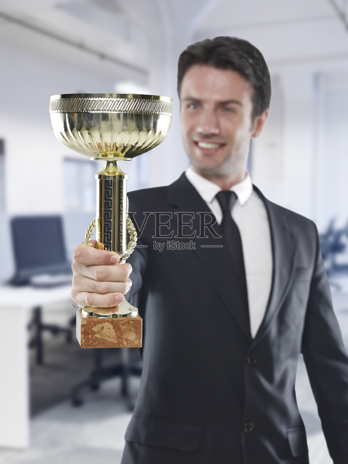 身着西装的快乐商人在现代办公室里举着金奖杯。照片摄影图片