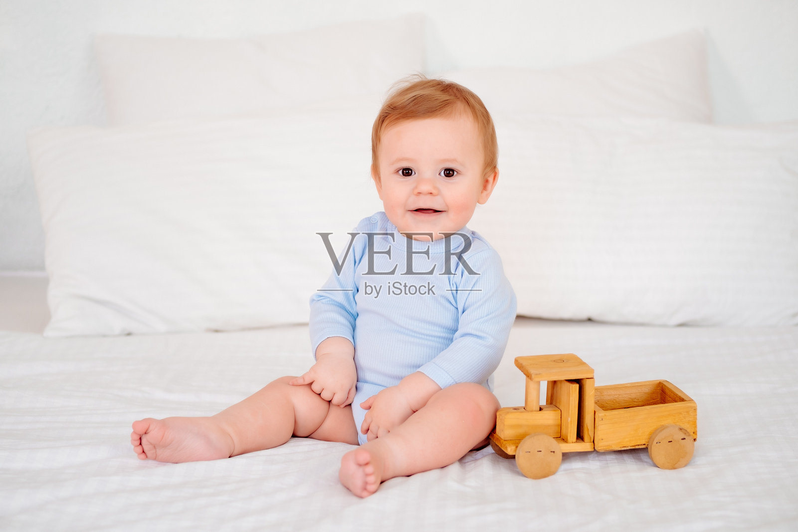 一个穿着蓝色连体衣的可爱小孩,在白色的床上玩着木制玩具车,脸上洋溢着快乐的笑容。照片摄影图片