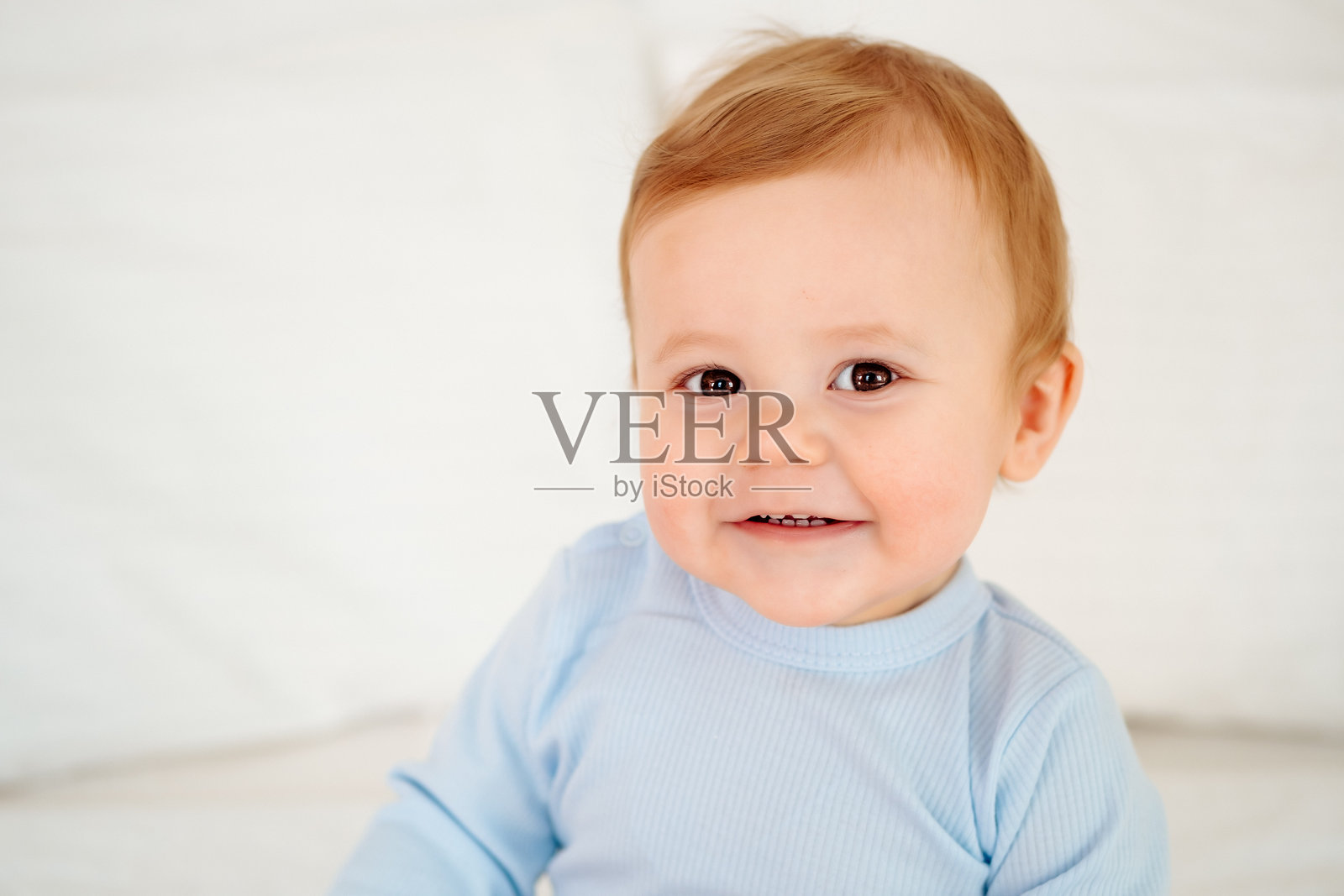 婴儿特写肖像,一个穿着蓝色连体衣的快乐微笑小宝宝在白色床上照片摄影图片