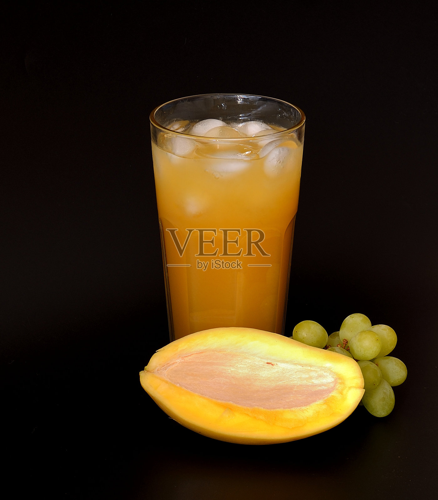 黑色背景上放着装有冰块的果汁混合饮料高杯,旁边有成熟的芒果和一串绿葡萄。照片摄影图片