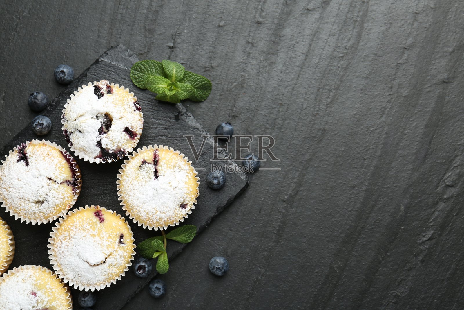 美味的蓝莓玛芬,撒有糖粉,用薄荷叶点缀,摆放在深色纹理桌面上,俯视角度拍摄,并留有文字空间。照片摄影图片