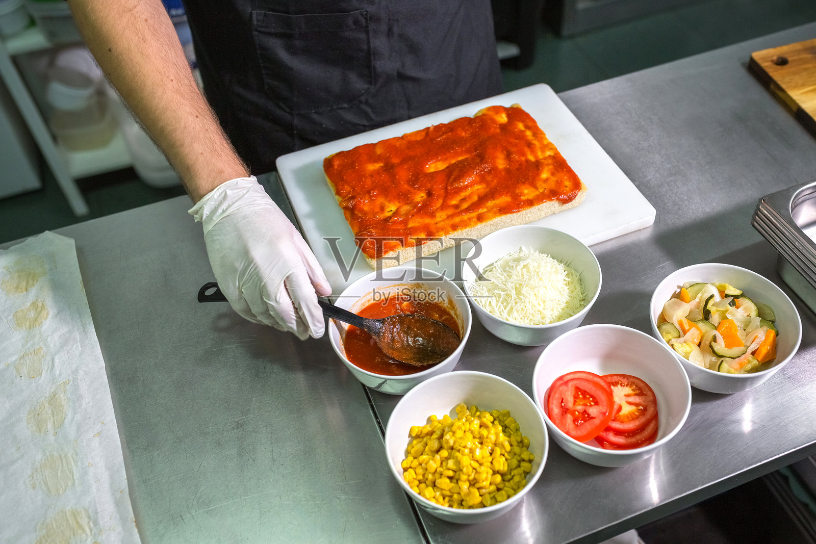在餐厅厨房里,一位戴着手套、身份不明的厨师正在舀取番茄酱,准备制作披萨。照片摄影图片