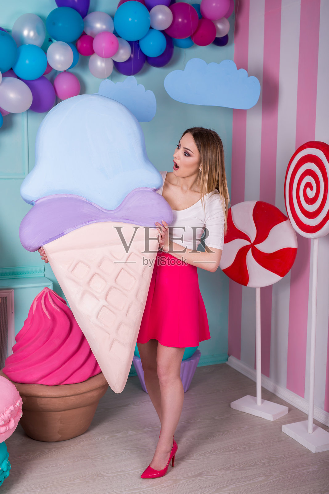身穿粉色连衣裙的年轻女子肖像,她手中拿着一个大冰淇淋,在装饰背景前摆姿势。令人惊叹的甜食爱好者,周围摆满了玩具糖果。照片摄影图片