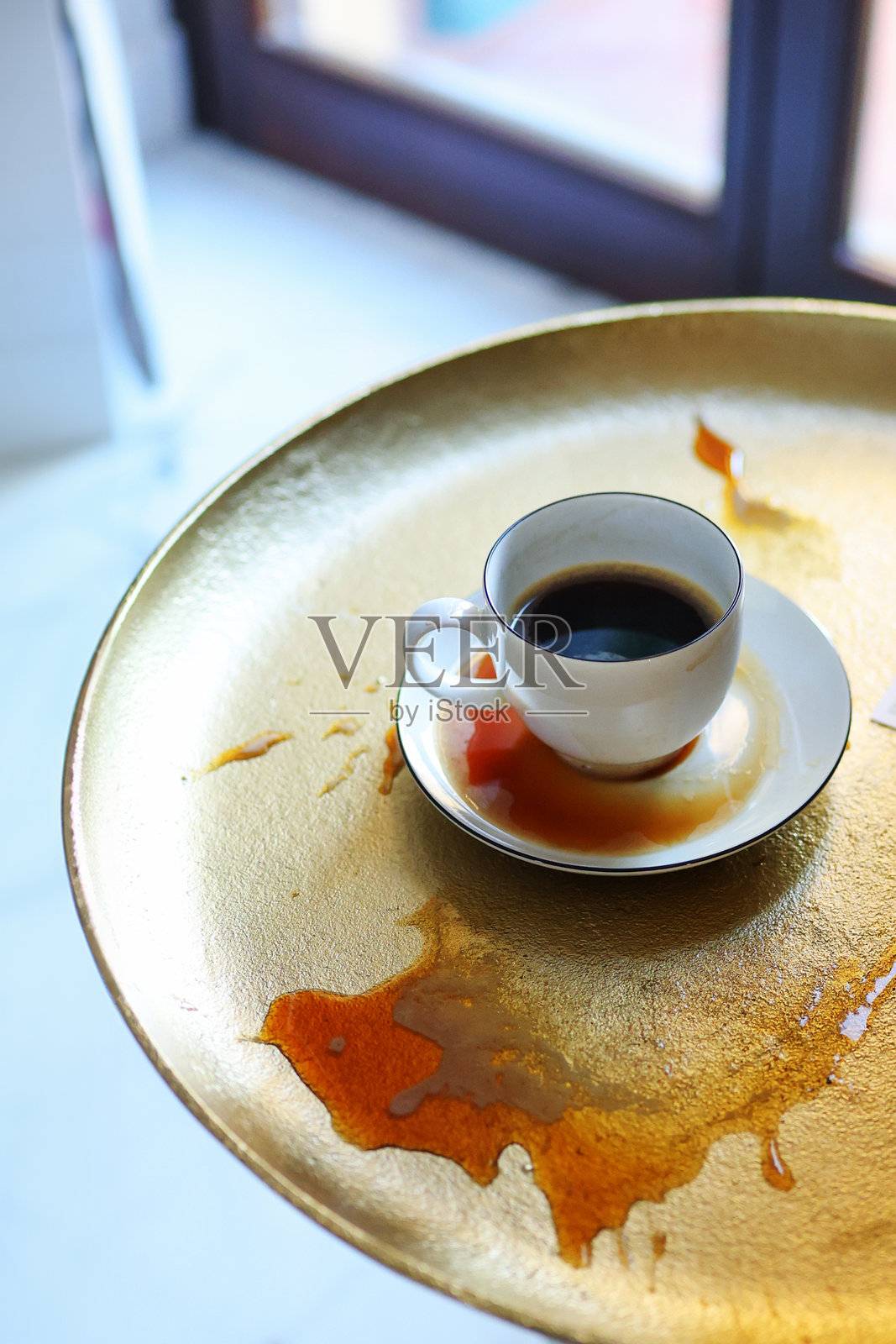 极简风格的静物画,金色桌子上放着一个黑色咖啡杯,溢出的咖啡形成了抽象的污渍。概念是关于不完美、晨间例行事务和咖啡馆氛围。照片摄影图片