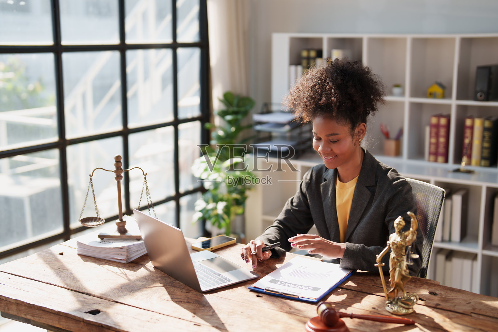 年轻女律师在办公室的木桌旁,手持法律合同,使用笔记本电脑工作,周围环绕着象征司法的天平和法槌等符号。照片摄影图片