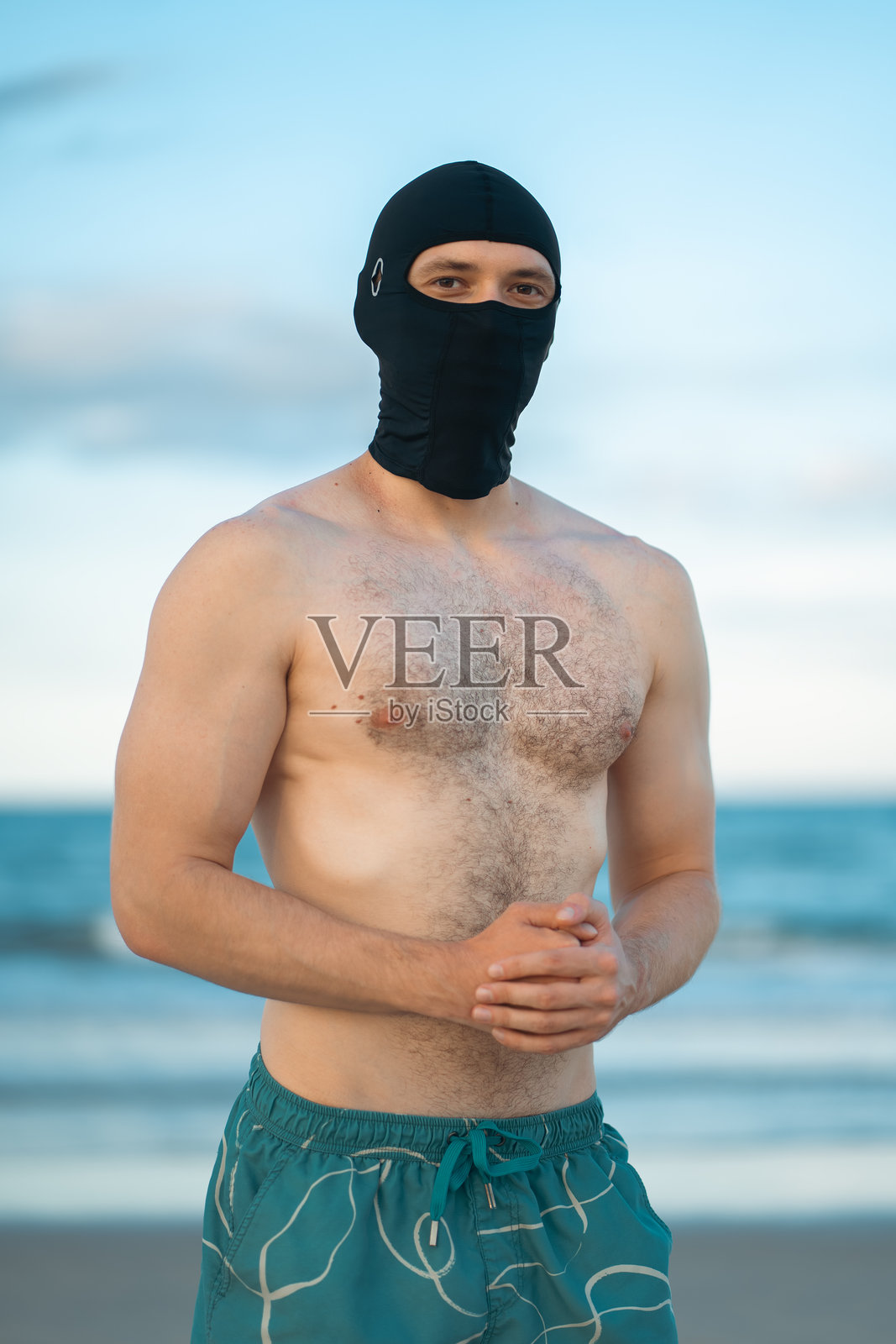 戴着口罩的白人男性,拥有健美的肌肉身材,站在阳光灿烂的日落海滩上,一位匿名的、有吸引力的男性运动员在海边或海岸线上行走。照片摄影图片