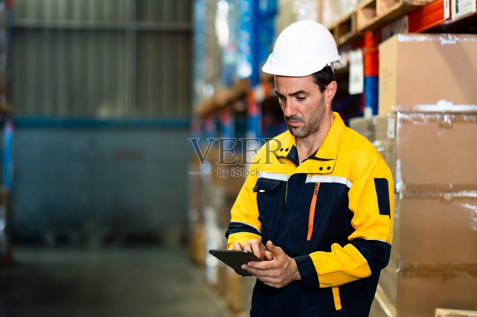 一名高加索男性物流工作人员,身着安全头盔和黄色防护服,在堆满货架的工业仓库中,手持平板电脑查看库存数据。照片摄影图片