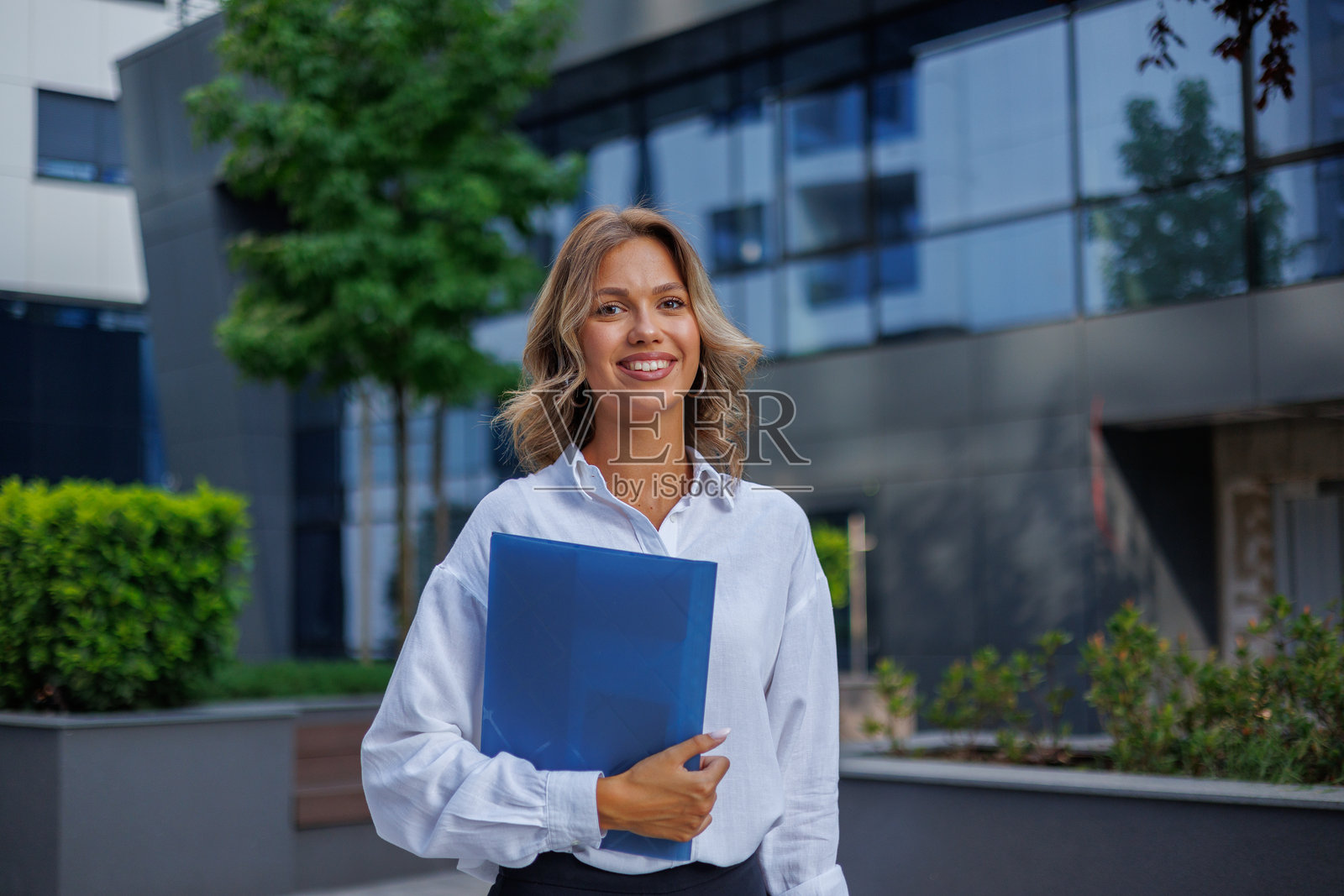 一位自信的职业女性,手持文件夹,面带微笑,站在现代化的公司大楼前照片摄影图片