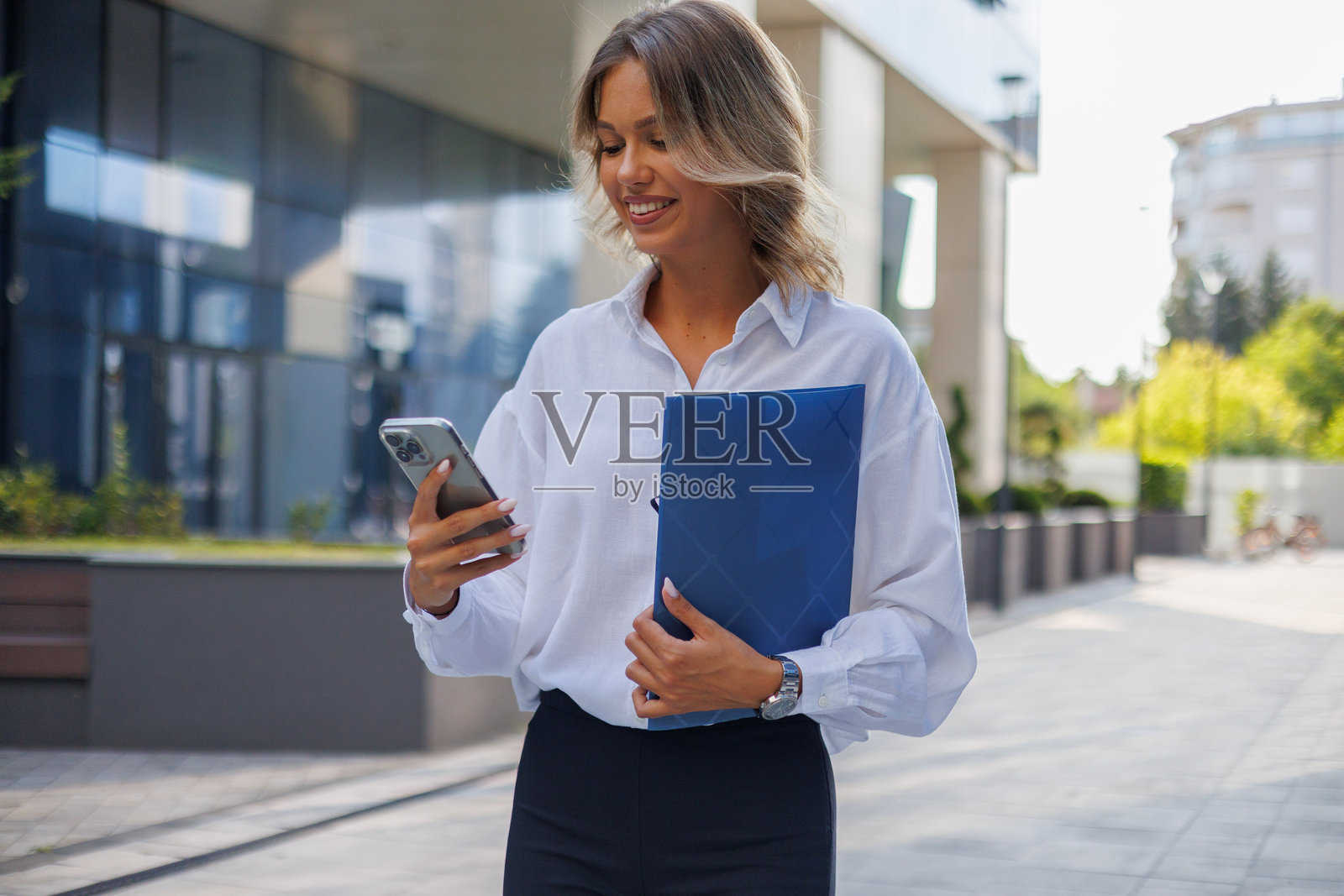 一位女商人正在户外使用智能手机并拿着文件袋,她面带微笑地查看信息。照片摄影图片