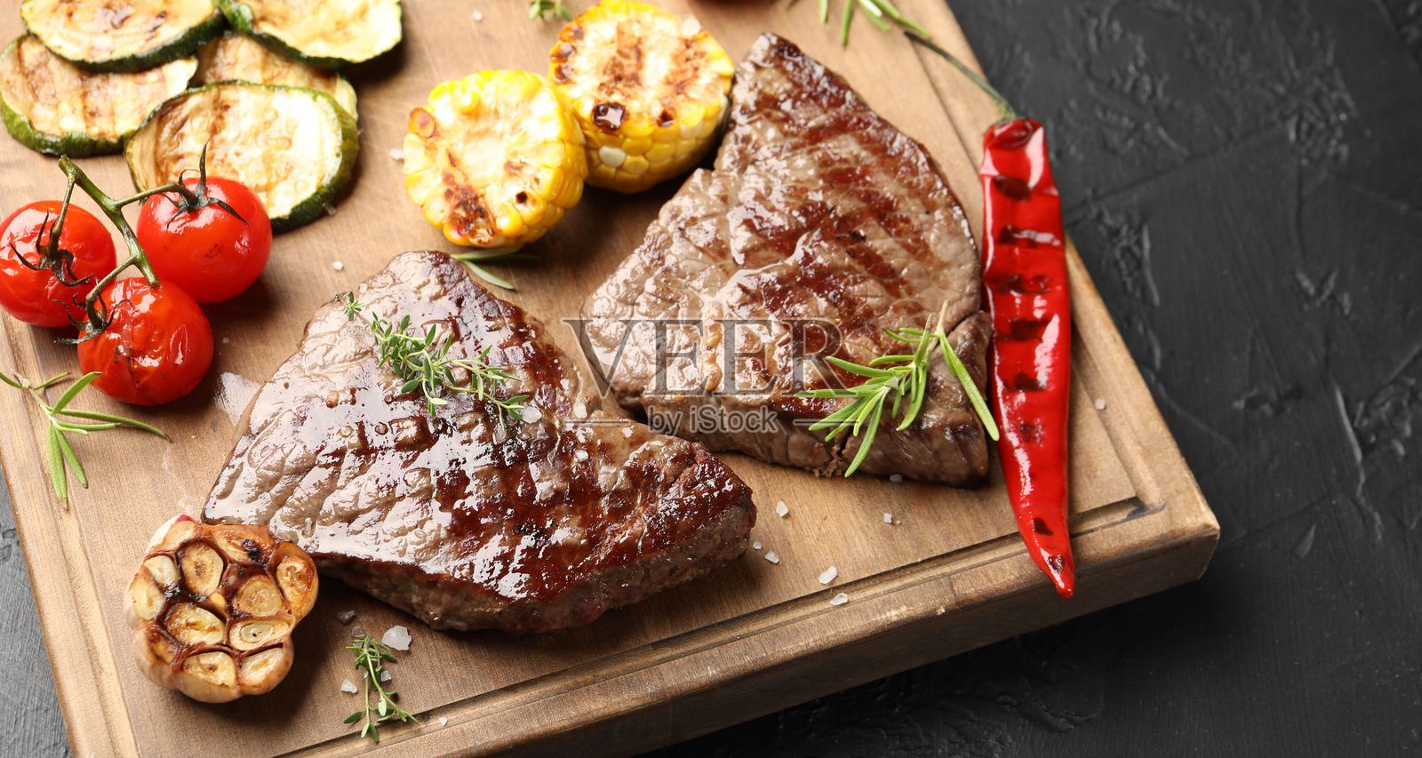 深色纹理桌面上摆放着美味的烤牛肉牛排,配以香草和蔬菜,特写镜头。照片摄影图片