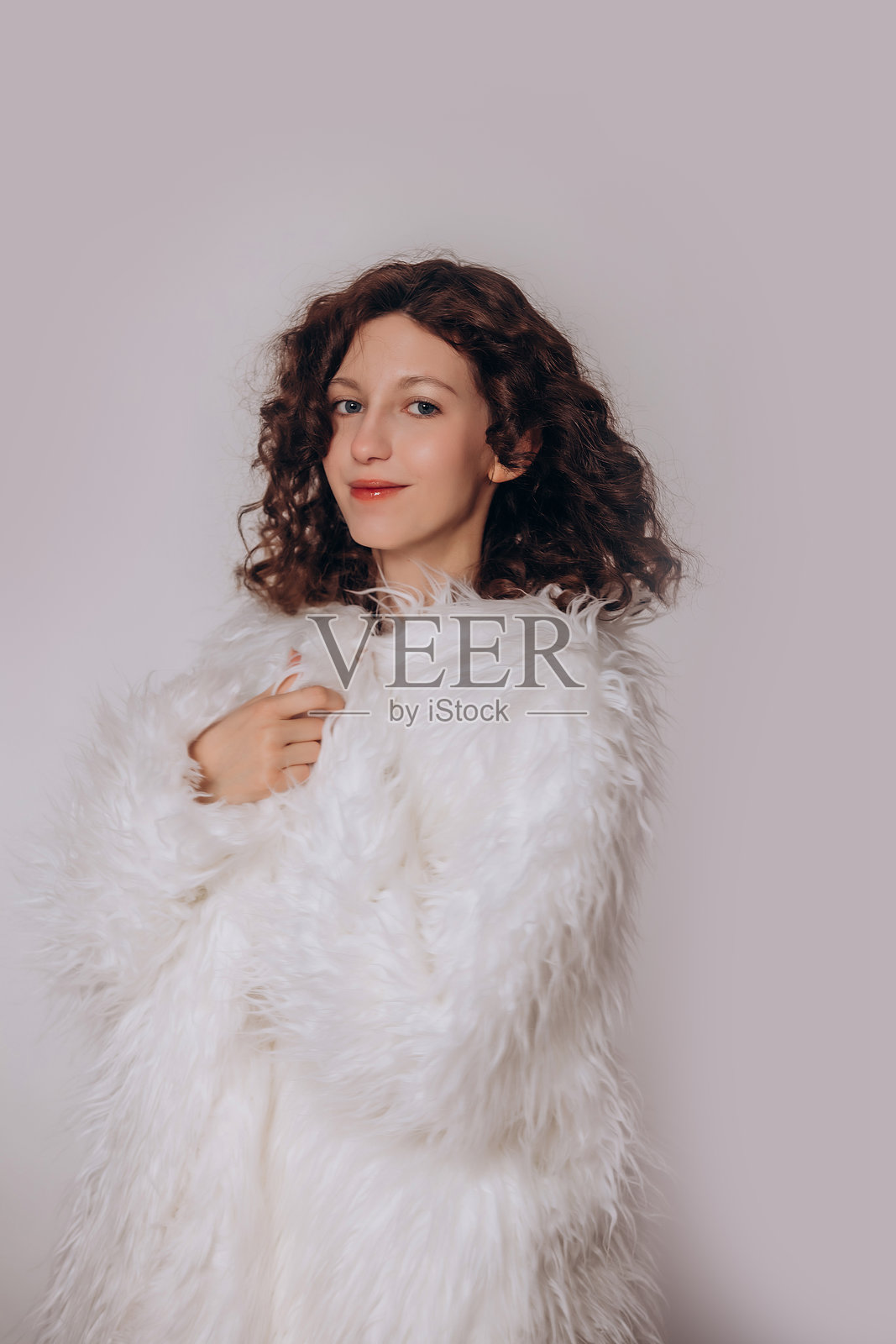 模特是一位年轻美丽的卷发棕发女孩,身穿白色蓬松皮草大衣,站在白色背景前。照片摄影图片