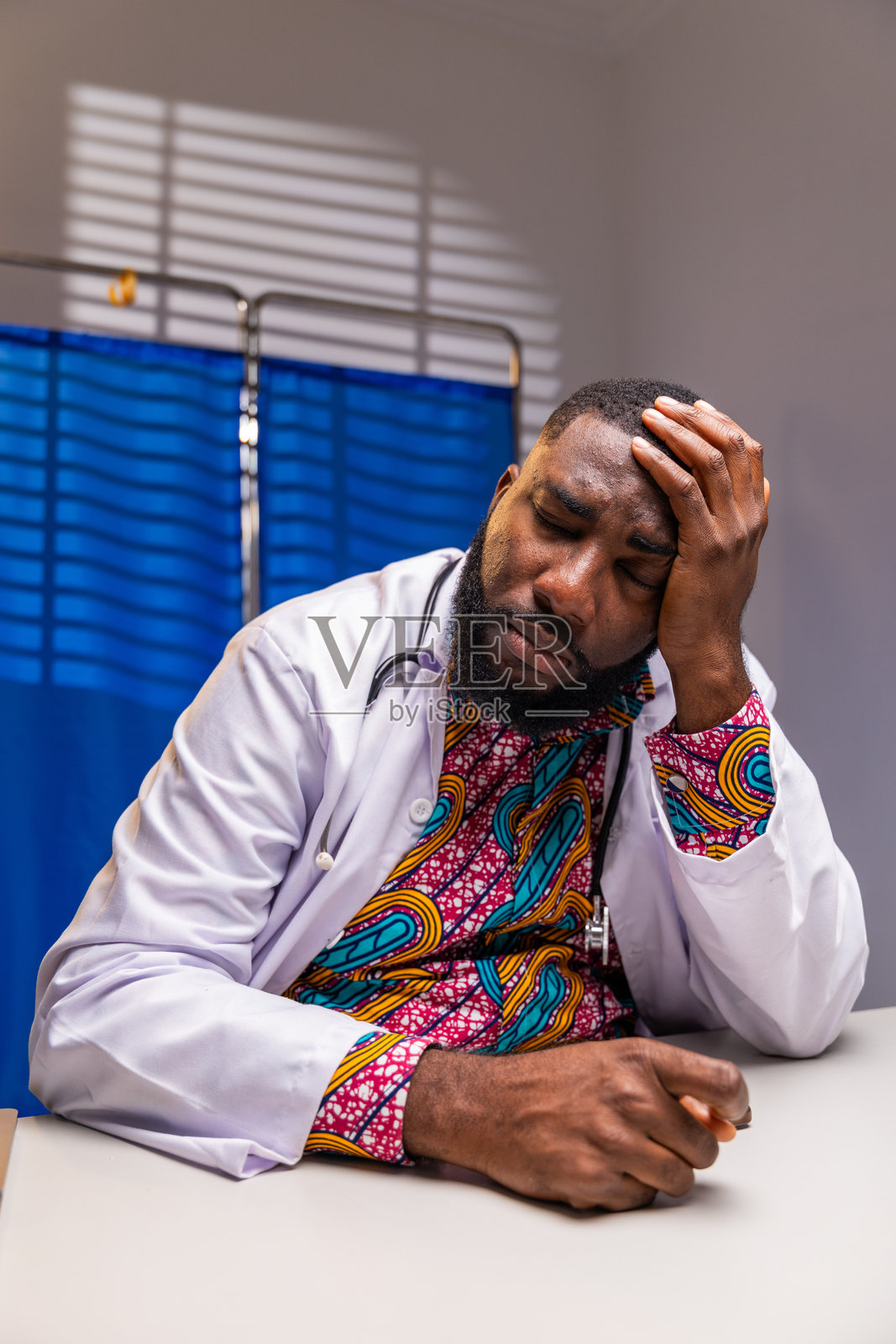 一名压力过大的非裔医生,闭着眼睛,双手放在太阳穴上,坐在医院的办公桌前,反映出职业倦怠和疲惫。照片摄影图片