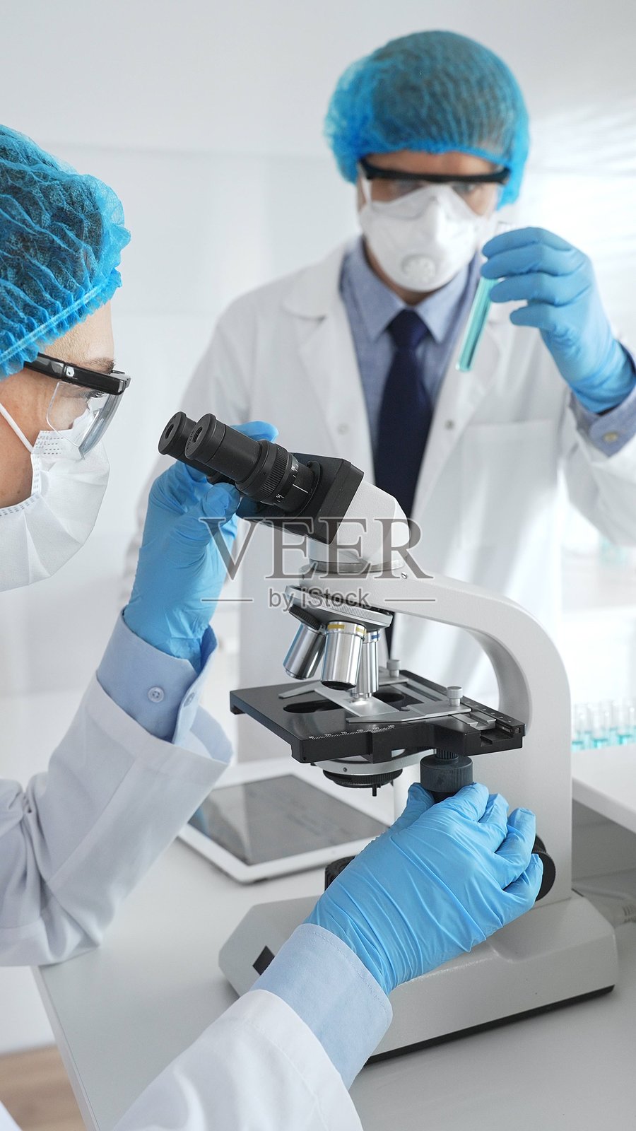 身穿实验服、戴着口罩和防护眼镜的两位科学家在实验室里用显微镜进行研究。医学与科学概念照片摄影图片