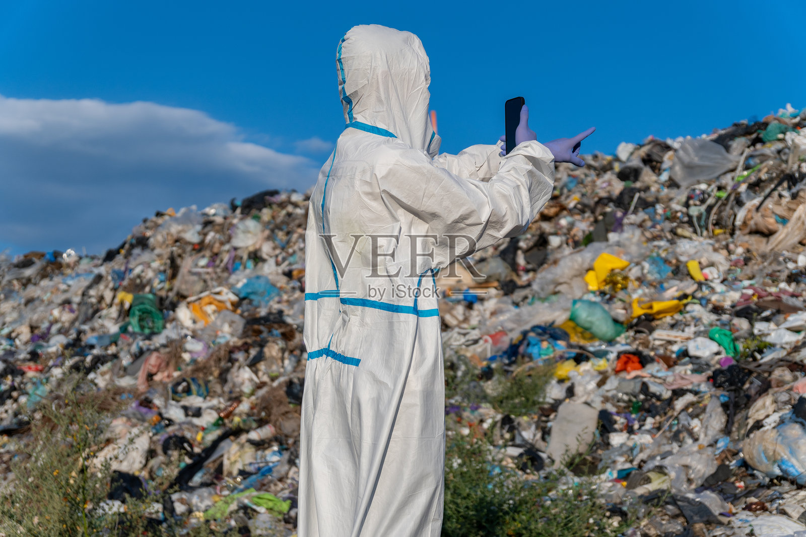 一名穿着防护服戴着手套的人,在一个垃圾填埋场的大堆垃圾上指指点点,背景是蓝天白云。照片摄影图片