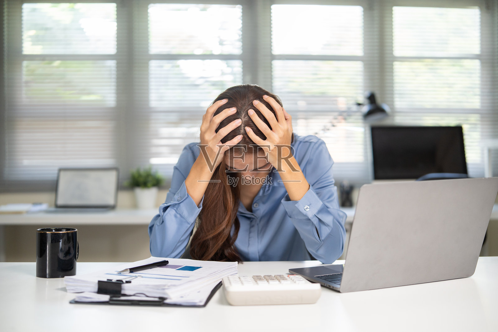 一位压力巨大的女商人在办公桌前用笔记本电脑工作,双手抱头,不堪重负。照片摄影图片