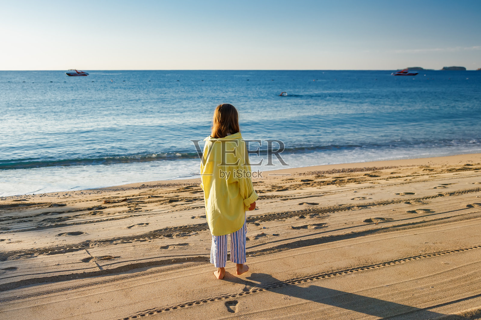 一名穿着黄色上衣和条纹裤子的年轻女孩,赤脚走在沙滩上,在日出时望着蔚蓝的大海,象征着夏日假期和宁静的旅行概念照片摄影图片