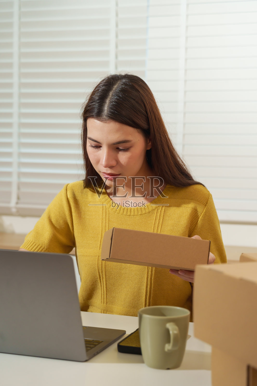 亚洲女企业家在家中办公室,手持纸箱,用笔记本电脑查看在线订单,展现出专注和专业的电子商务小企业包装管理能力照片摄影图片