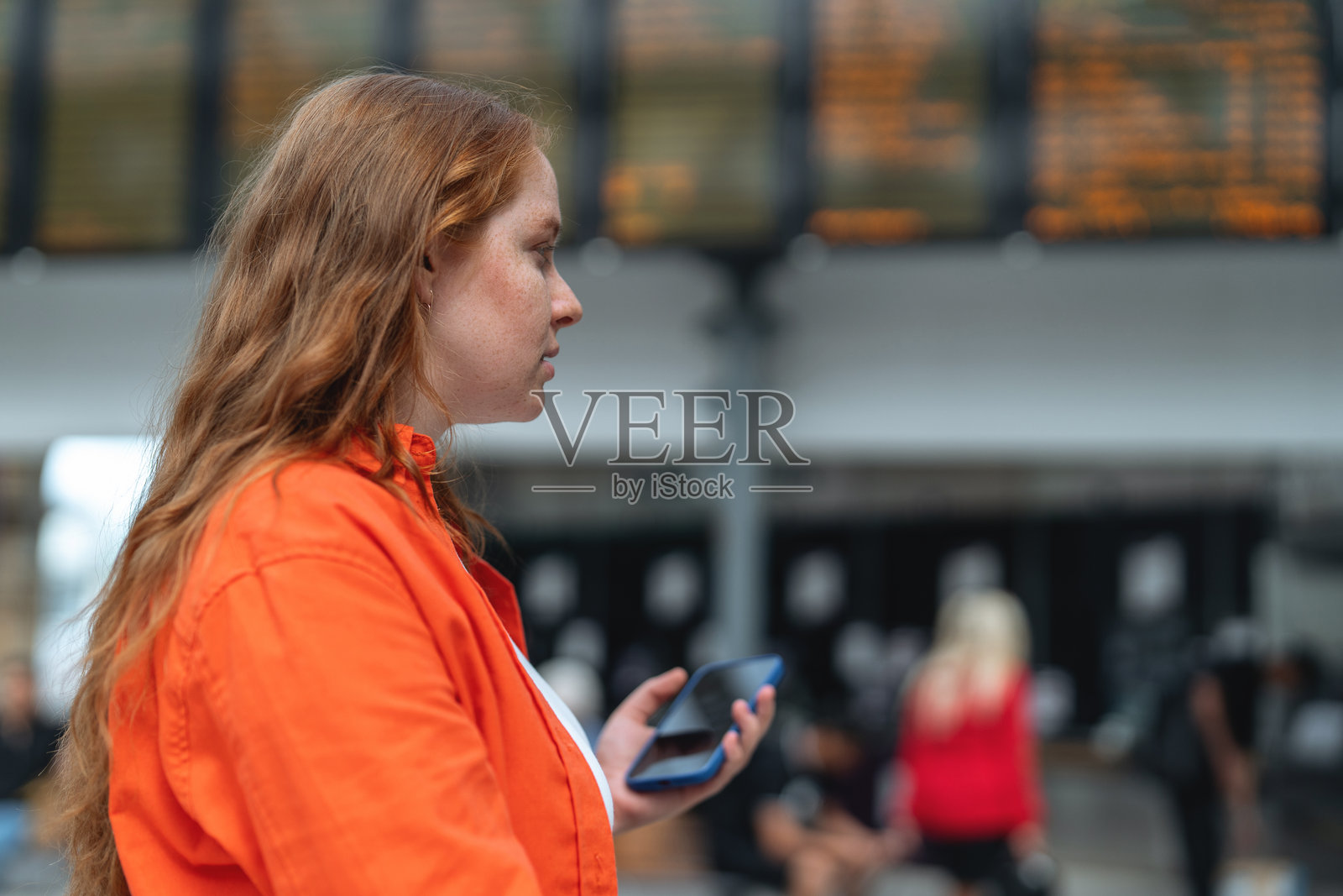 一位女士在繁忙的白天于交通枢纽站候车,手中拿着手机。照片摄影图片