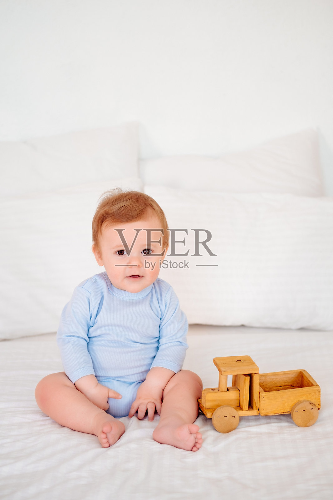 一个穿着蓝色连体衣的快乐微笑小宝宝在白色床铺上玩着木制玩具车照片摄影图片