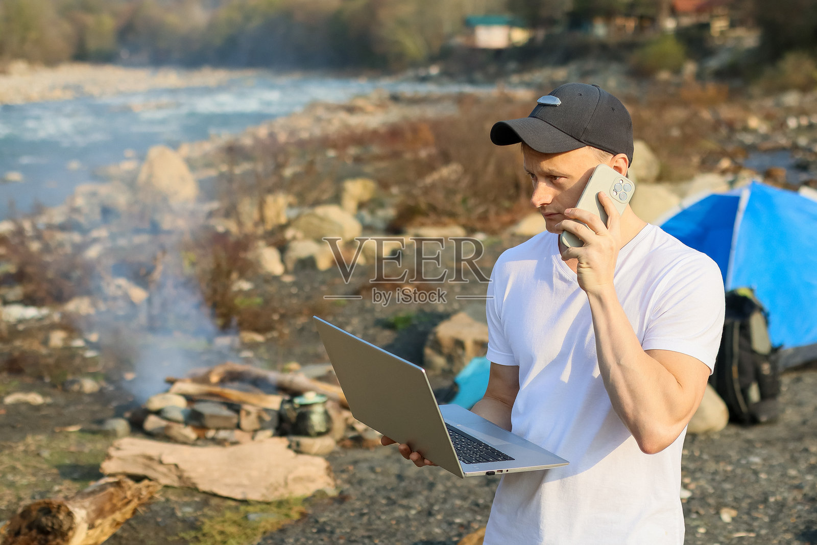男人在河边的户外营地里,一边打着电话,一边拿着笔记本电脑,旁边还有帐篷。照片摄影图片