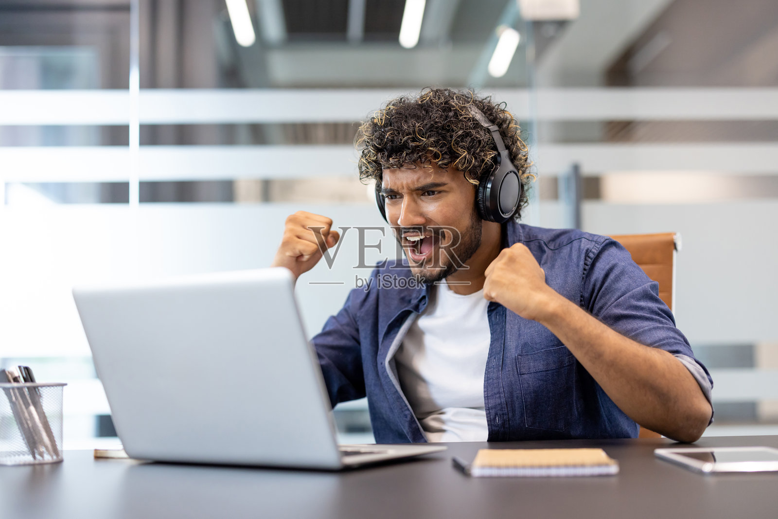 一位戴着耳机、看着笔记本电脑屏幕、欢欣鼓舞的年轻印度男子坐在办公室的桌前。照片摄影图片