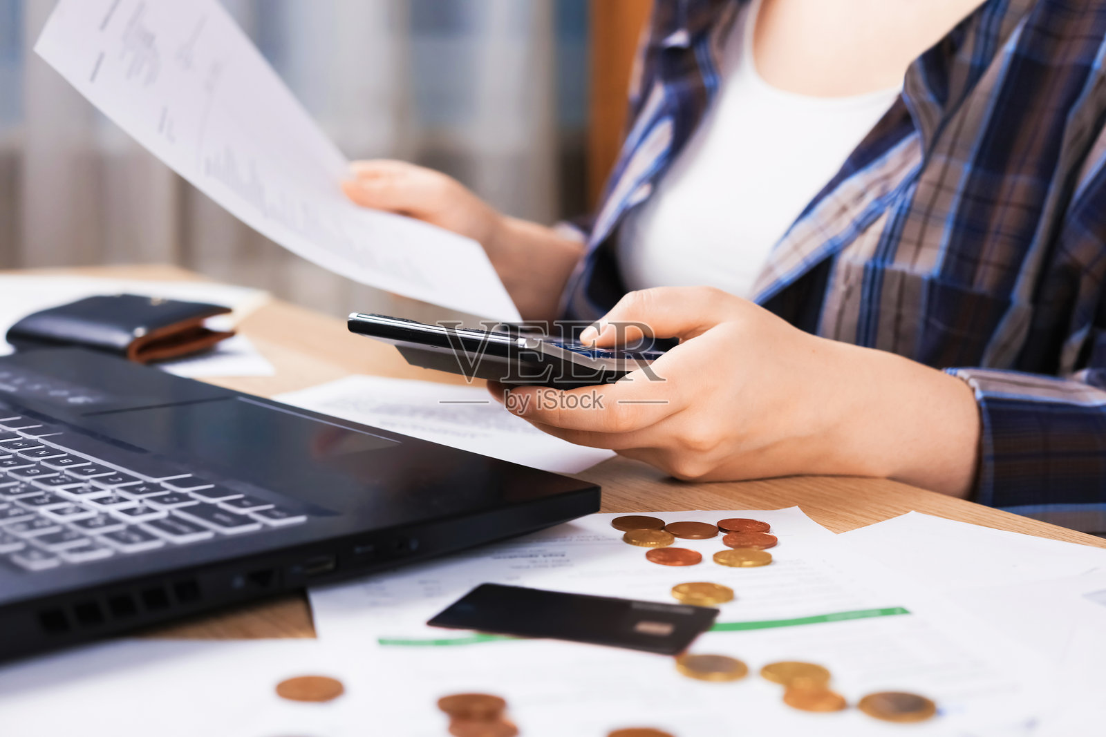 一位女士拿着账单,使用计算器,旁边放着一台笔记本电脑。桌上摆放着硬币和信用卡,体现了财务规划和家庭预算的概念。照片摄影图片