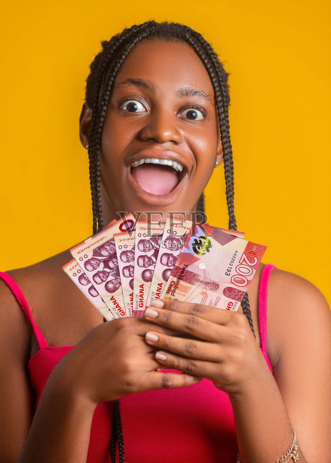 一位年轻女子身穿粉色上衣,扎着辫子,手持一扇加纳200塞地纸币,在明亮的黄色背景前,她张大嘴巴,脸上洋溢着惊喜和喜悦之情,表情十分夸张。照片摄影图片