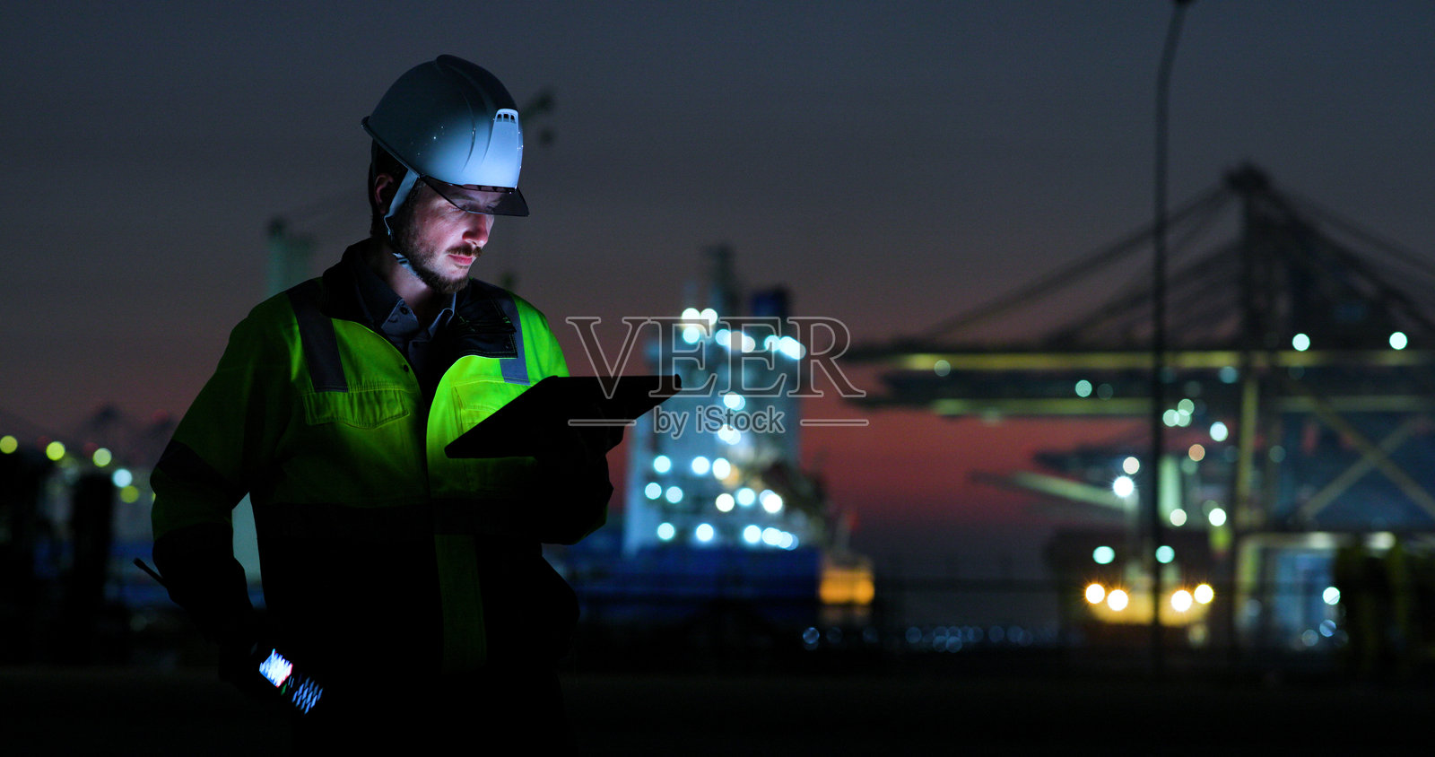 一位身穿头盔和高可见度夹克的港口安全官,在夜间繁忙的港口运营中,一边通过无线电沟通,一边拿着平板电脑。照片摄影图片