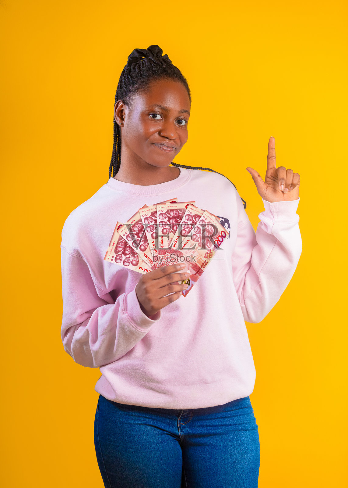 一位留着编织发型、身穿粉色运动衫的年轻非洲黑人女性,站在鲜艳的黄色背景前。她一手拿着一扇加纳200塞地纸币,另一只手向上指着,脸上带着自信而俏皮的表情。照片摄影图片