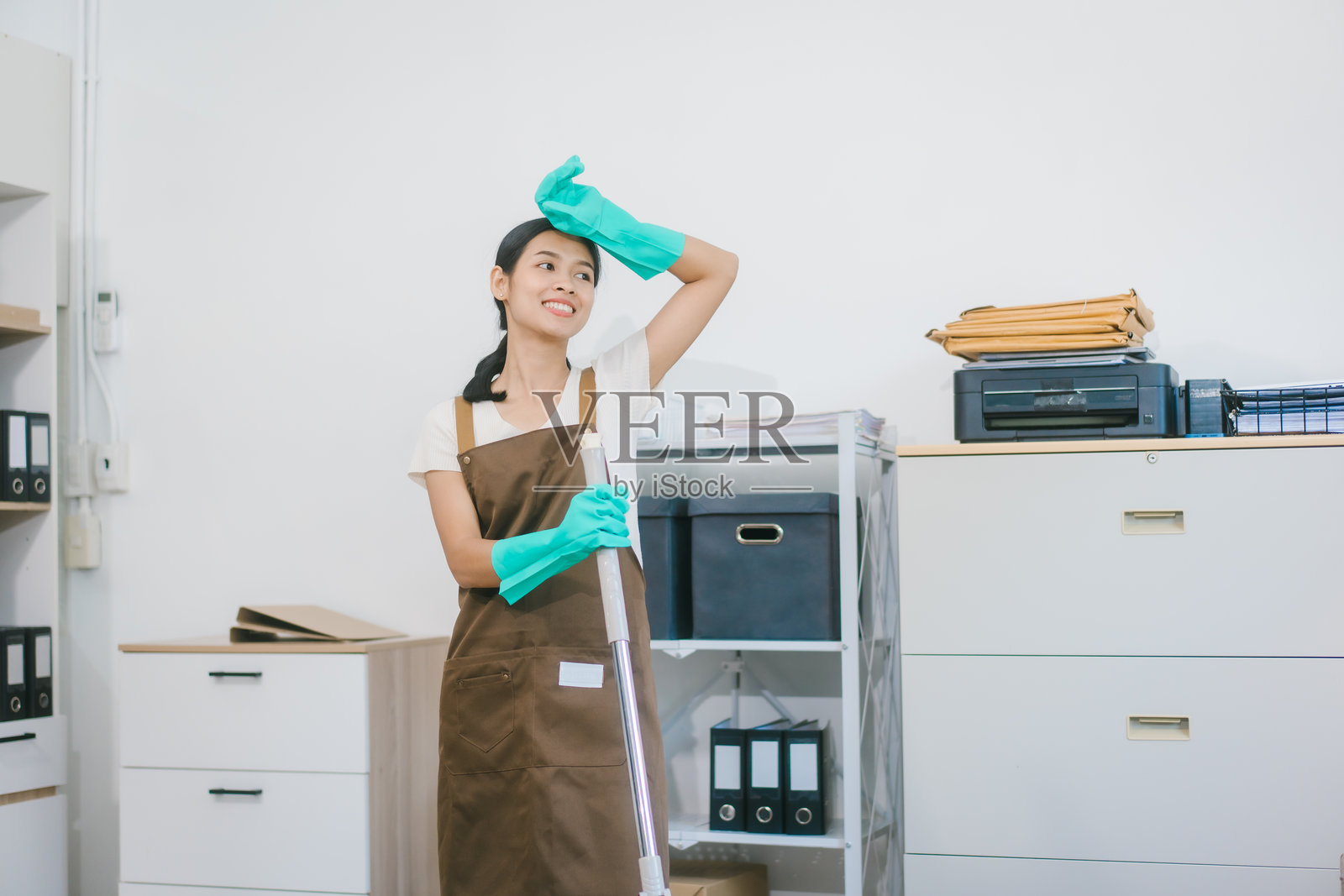一位穿着手套和围裙的亚洲女性家政服务人员正在清洁现代办公室或家庭。她勤奋工作,保持卫生和整洁,体现了专业的清洁服务。照片摄影图片
