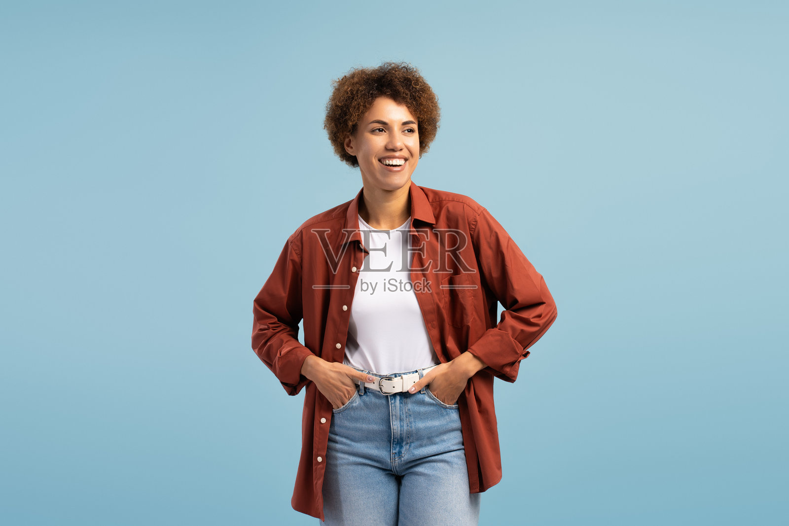 微笑着的非裔美国女性双手插兜,在蓝色背景前摆姿势照片摄影图片